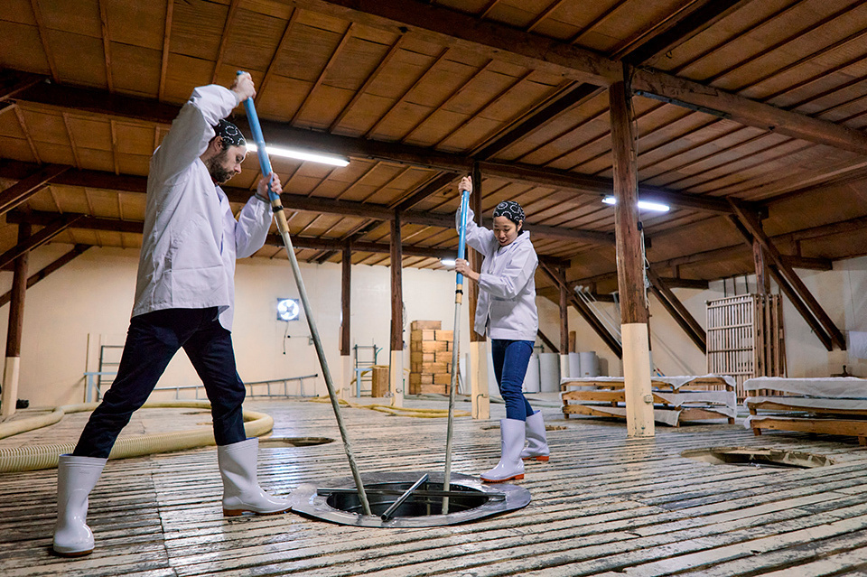 Two people in sake brewery stirring fermentation tank.