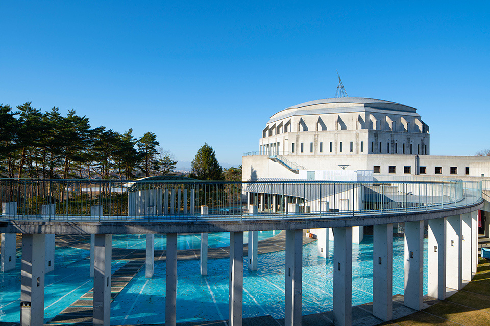 The Octagonal White Building is located behind a row of white pillars in a circle and a blue floor front plaza in Shichigahama International Village.