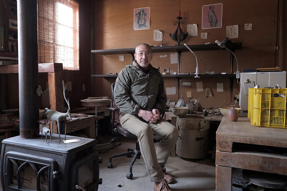 A man sitting in a rustic workshop filled with various tools and a wood-burning stove.