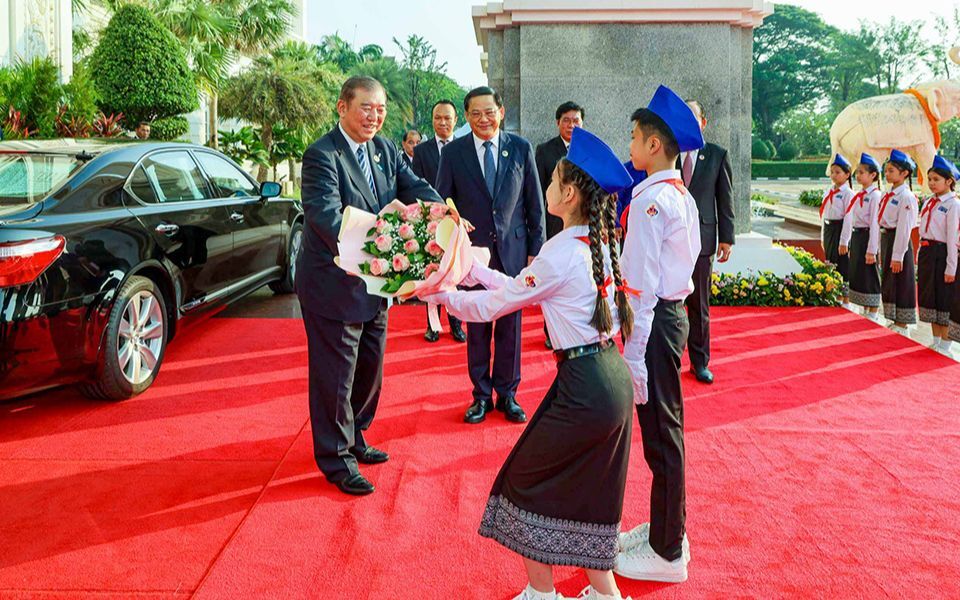 Prime Minister Ishiba receives a bouquet from two children in traditional uniforms on a red carpet, at a welcome ceremony in Lao PDR on October 11, 2024.