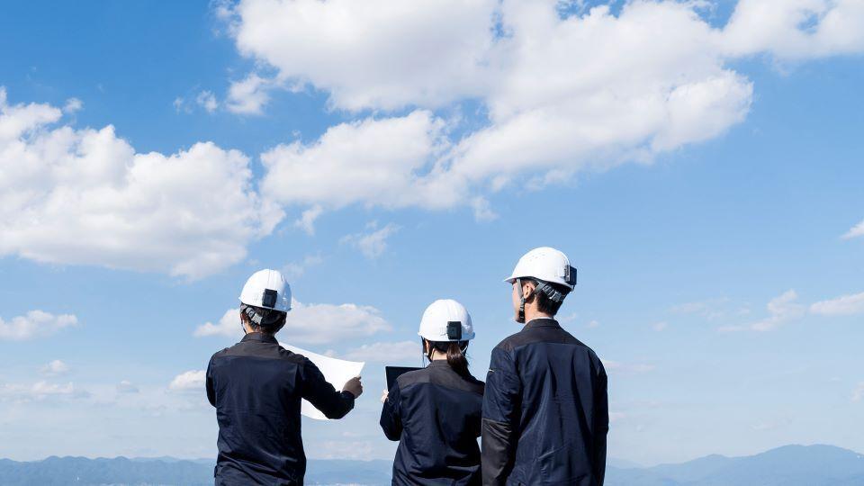 Three people wearing white helmets are positioned on a hill, observing something off in the distance.