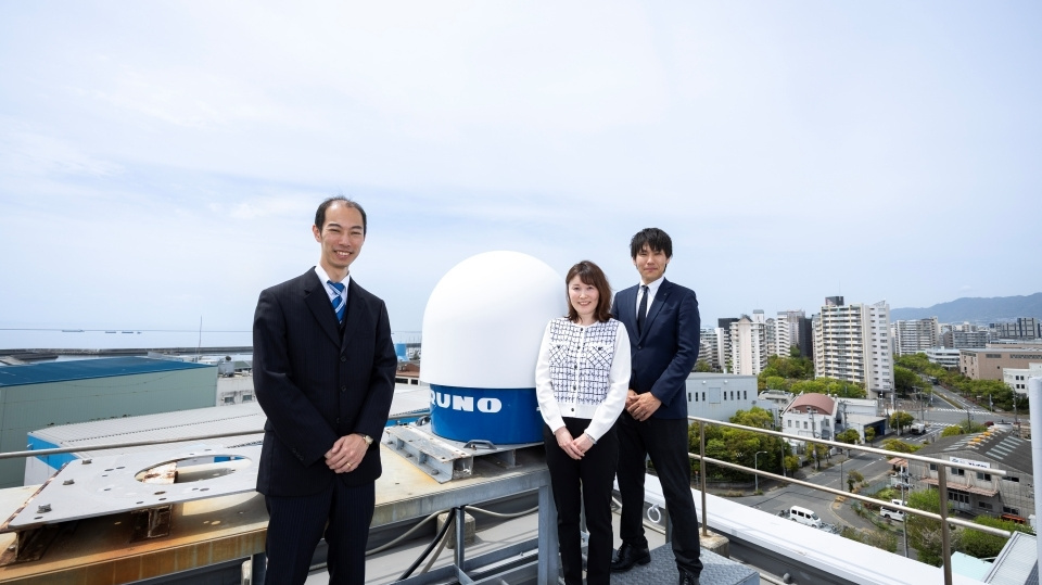 Three people standing on a rooftop in front of a blue and white compact weather radar system