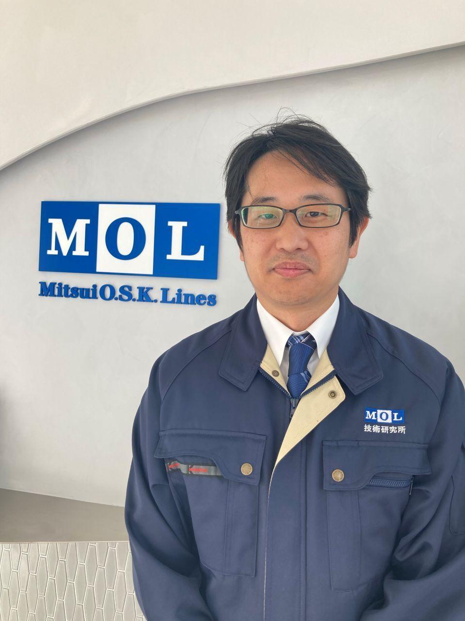 A Japanese man in a blue jacket and tie posing in front of the Mitsui O.S.K. Lines logo