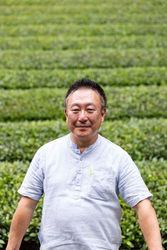 A man in a grey shirt posing in front of an orderly tea field