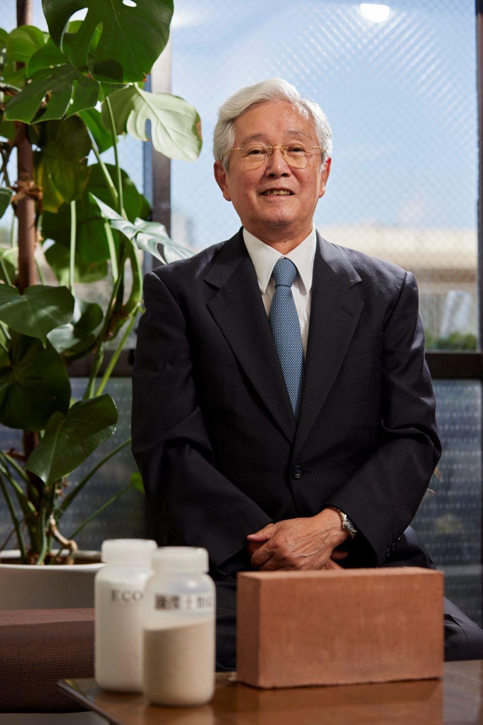 A white-haired Japanese man in a dark suit, standing behind a large brick and bottles of white powder