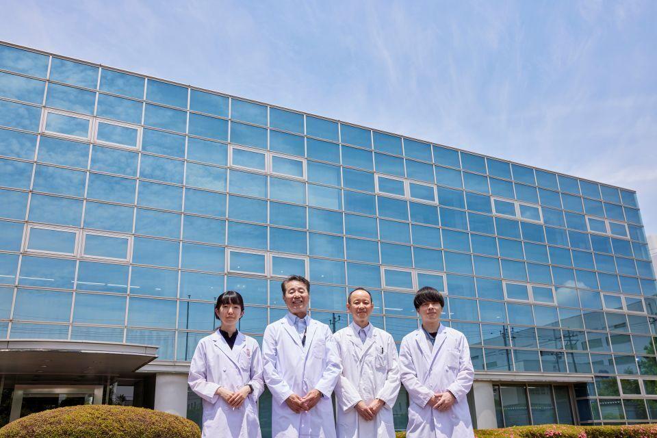 A team of four scientists in white lab coats posing in front of a modern, glass-fronted building